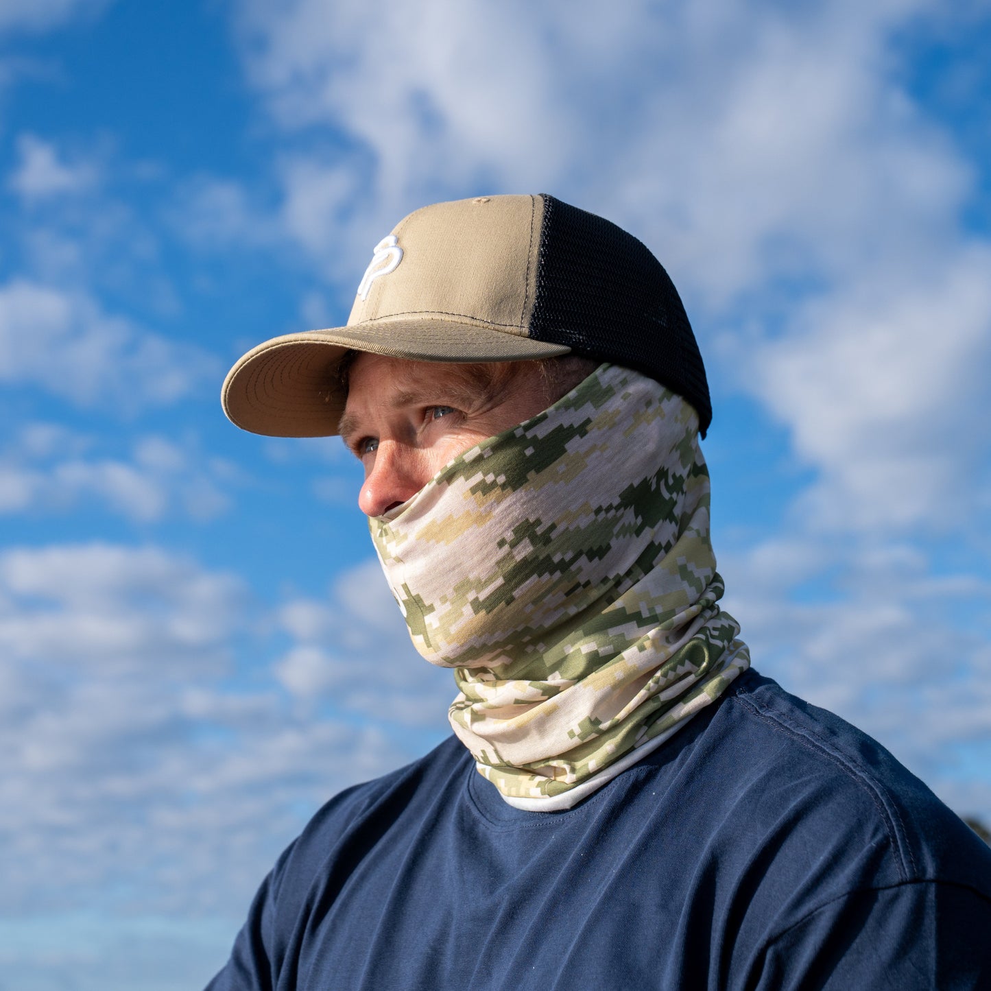 Person wearing a cap and camouflage face mask against a blue sky with clouds